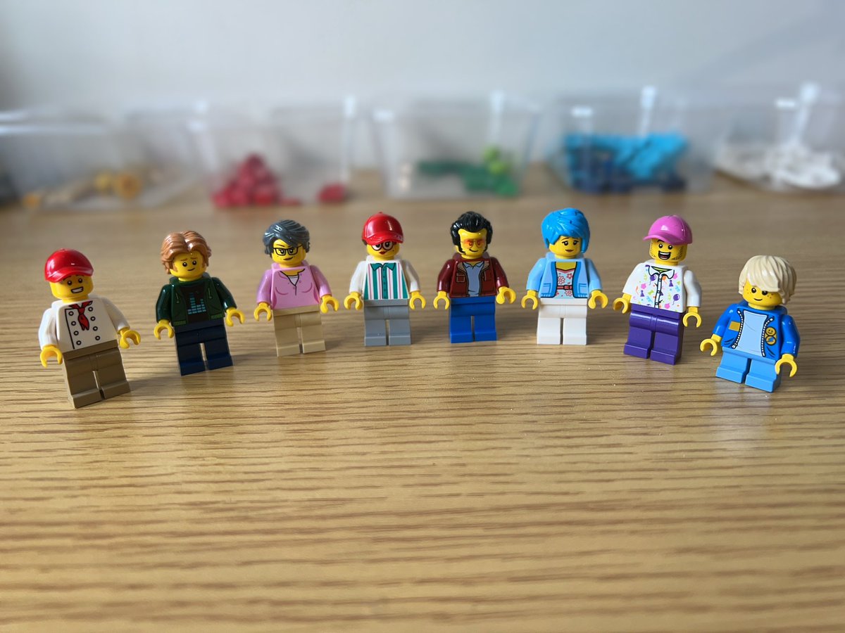 Eight lego minifigures standing side by side. There’s a person in a chef’s jacket and red baseball cap, a person in a green hoodie, a lady in a pink blouse, a woman in a blue and white striped shirt, a man in a red leather jacket, a person with blue hair and a blue cardigan, a person in a white shirt with pictures of balloons, and a child with a denim jacket.