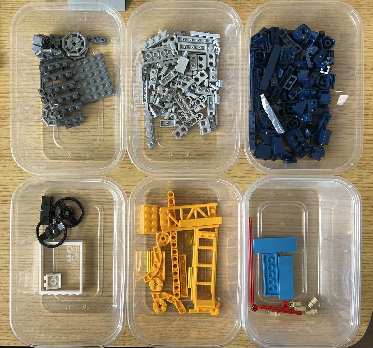 Six tubs of lego sorted by colour. Most of the pieces are grey or dark blue, but there are some yellow roller coaster track pieces and three light blue tile pieces.